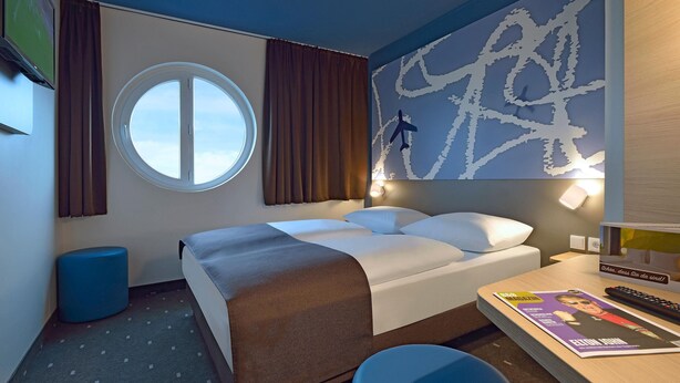 A hotel room with a bed, a round window, a wall art piece, a desk with a remote control, and a magazine. - B&B Hotel Frankfurt-hahn Airport (Lautzenhausen)