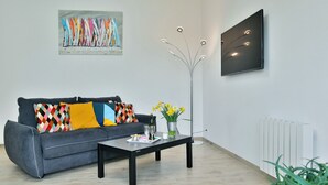 Un salon moderne avec un canapé gris, des coussins colorés, une table basse avec des boissons et un vase de fleurs, un lampadaire et un tableau au mur.