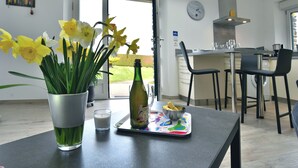 Une cuisine moderne avec un coin repas, comprenant une table avec une bouteille et un bol de chips, un vase avec des fleurs et une fenêtre donnant sur un paysage verdoyant.