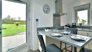 Une cuisine moderne avec une table à manger pour deux personnes, une horloge au mur et une vue sur le jardin par la fenêtre.