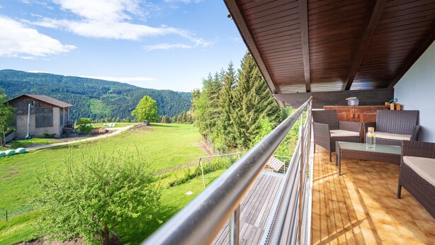 A balcony with wicker furniture, a glass table, and a view of a green landscape with trees and a house in the background.