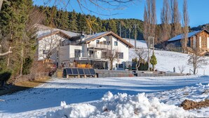 A modern two-story house with a balcony, surrounded by a snowy landscape and a forested hill.