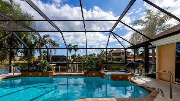 A pool area with a glass enclosure, surrounded by palm trees and a view of residential buildings. - Summer Breeze - Gulf Access Location - Pool & Hot (Cape Coral)