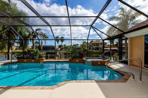 A pool area with a glass enclosure, surrounded by palm trees and a view of residential buildings. - Summer Breeze - Gulf Access Location - Pool & Hot (Cape Coral)