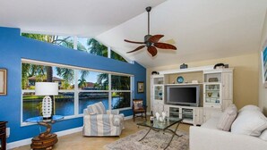 A living room with a large window, a ceiling fan, a TV, and a sofa. - Summer Breeze - Gulf Access Location - Pool & Hot (Cape Coral)