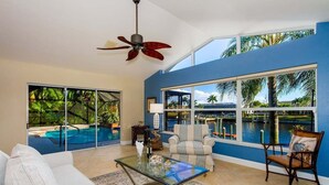 A spacious living room with a large glass coffee table, a sofa, and a chair. There is a ceiling fan, large windows with a view of a pool, and a blue accent wall. - Summer Breeze - Gulf Access Location - Pool & Hot (Cape Coral)