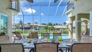 Comedor con cerramiento de vidrio, asientos al aire libre y vistas a una piscina y palmeras.
