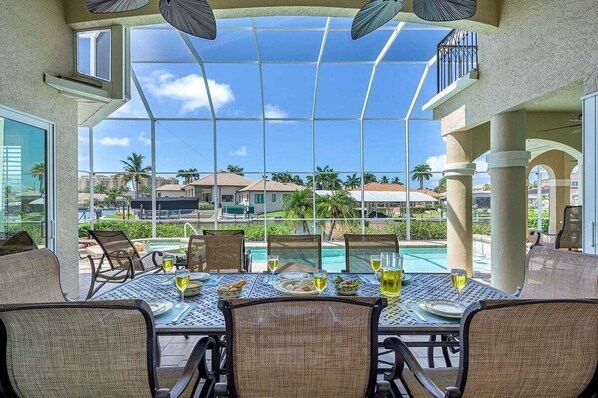 A dining area with a glass enclosure, outdoor seating, and a view of a swimming pool and palm trees.