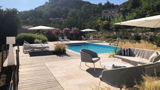 Un área de piscina con sillones de descanso y sombrillas, rodeada de follaje y colinas.