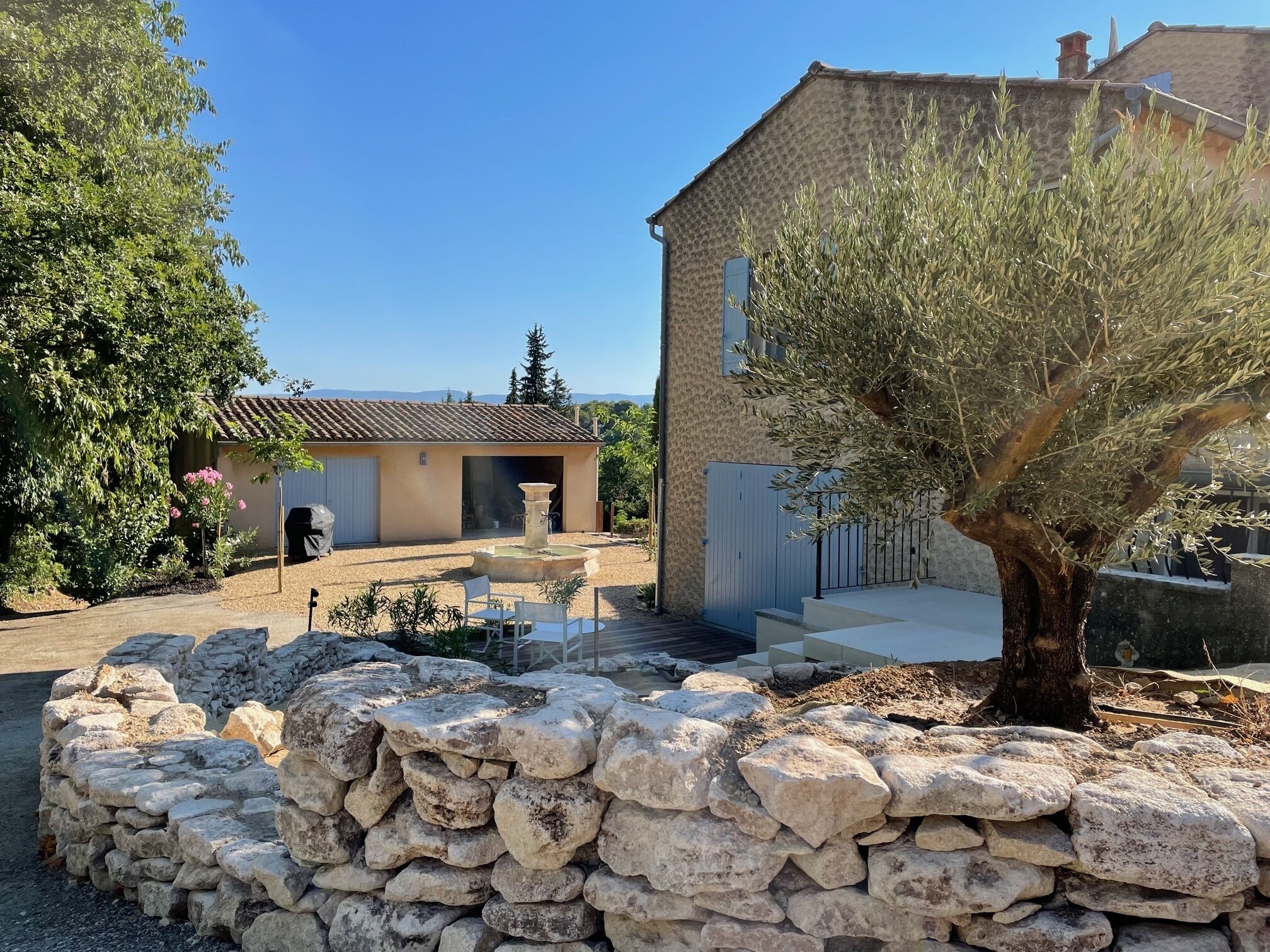 Un patio con paredes de piedra, un edificio rústico, mesas al aire libre y un gran árbol.
