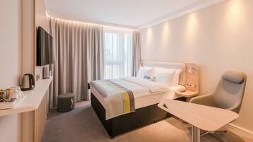 Una habitación de hotel con cama, escritorio, silla, televisor y ventana con cortinas.