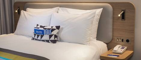 A hotel room with a bed, pillows, a bedside table with a phone, and a wall-mounted light.