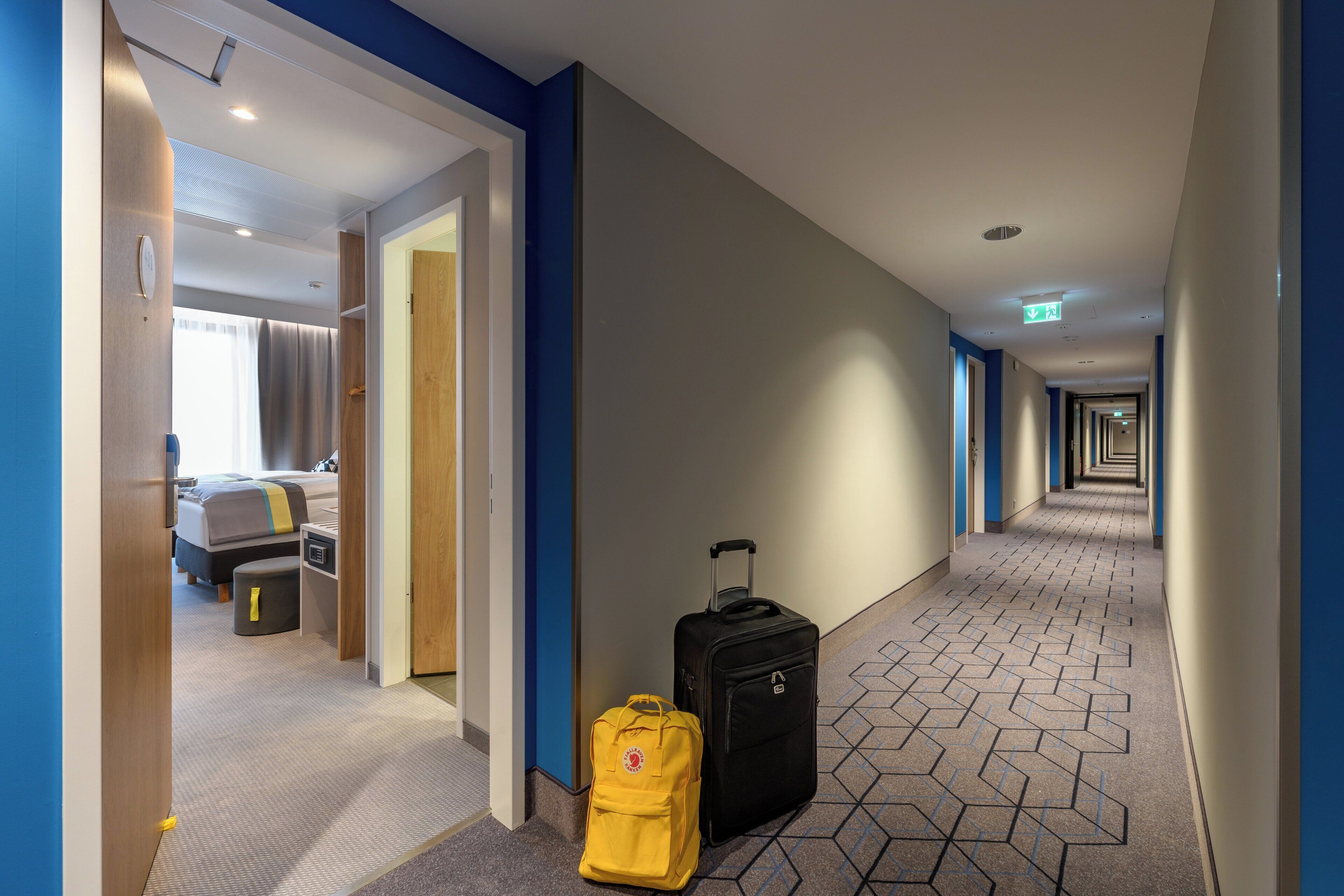 A hotel hallway with a bed, a suitcase, and a yellow bag.