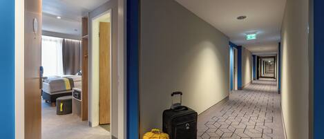 A hotel hallway with a bed, a suitcase, and a yellow bag.