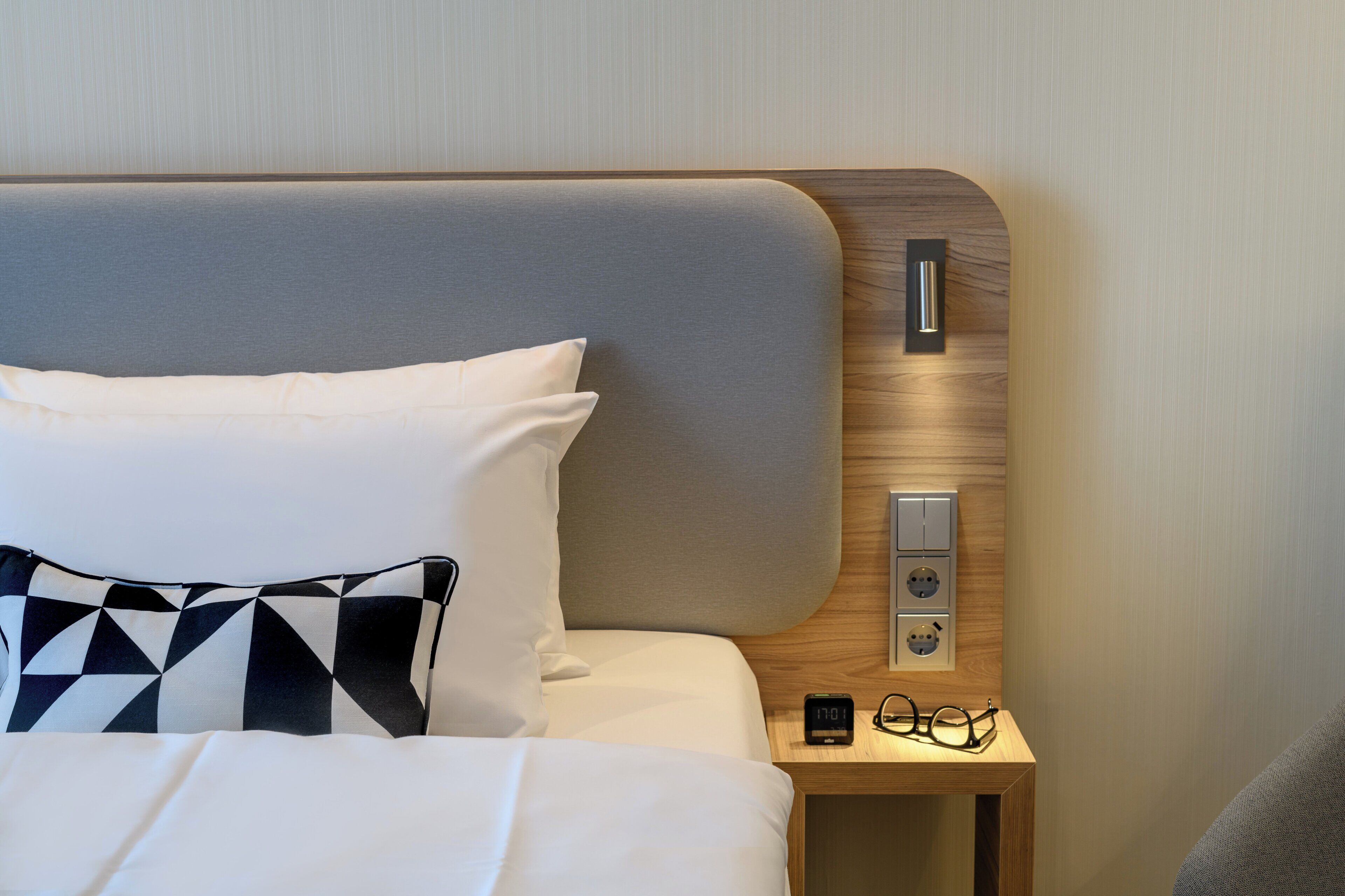 A neatly made bed with white and black pillows, a wooden headboard, and a bedside table with a digital clock and glasses.