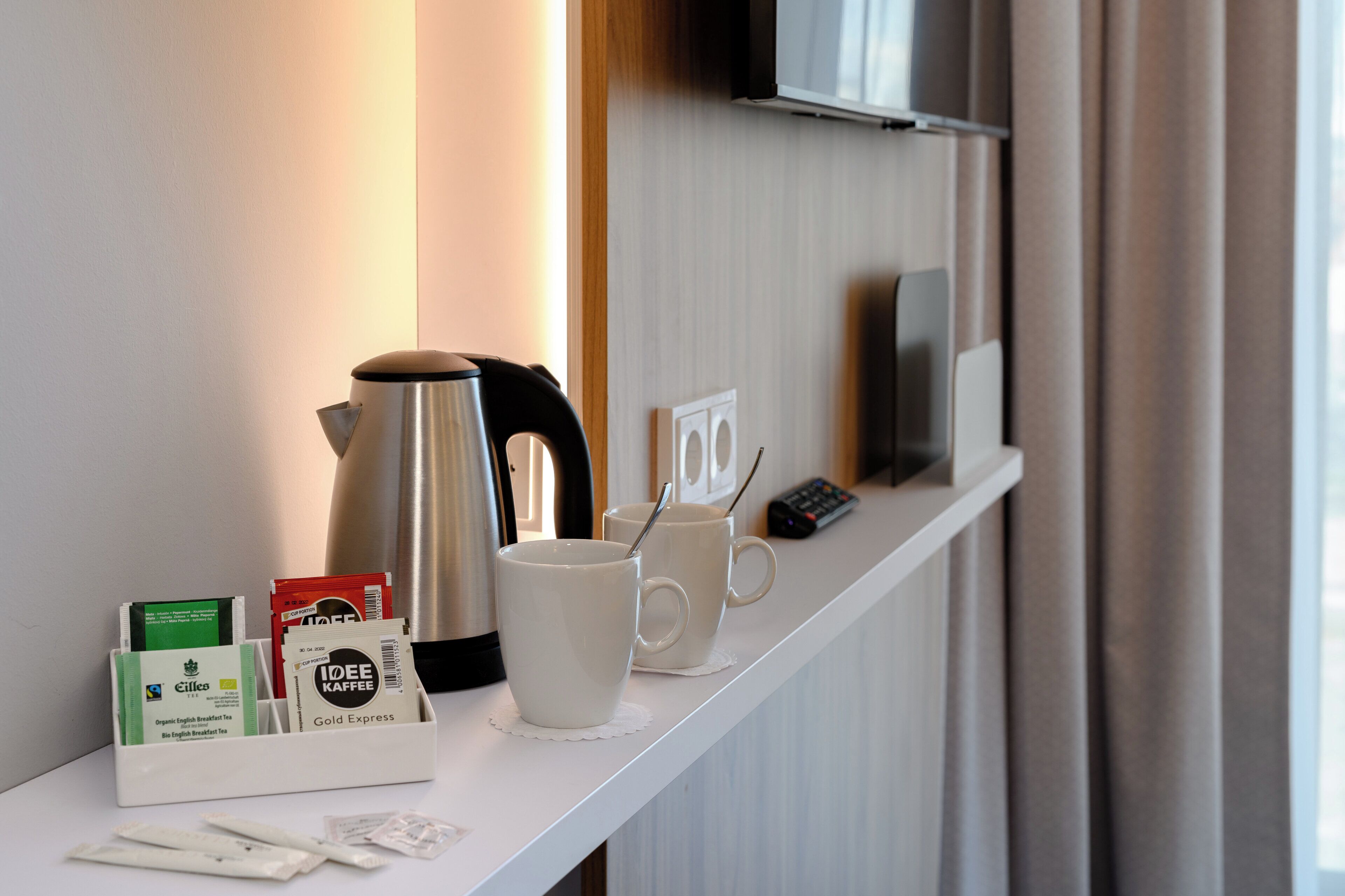 Área de desayuno en una habitación de hotel, con una cafetera, dos tazas, un control remoto y una caja de bolsitas de té.