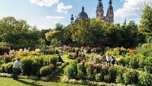 A garden with manicured lawns and flower beds, a historic building with two towers in the background, and people walking and standing in the garden. - Holiday Inn Express Fulda by IHG (Fulda)
