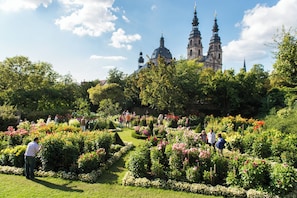A garden with manicured lawns and flower beds, a historic building with two towers in the background, and people walking and standing in the garden.