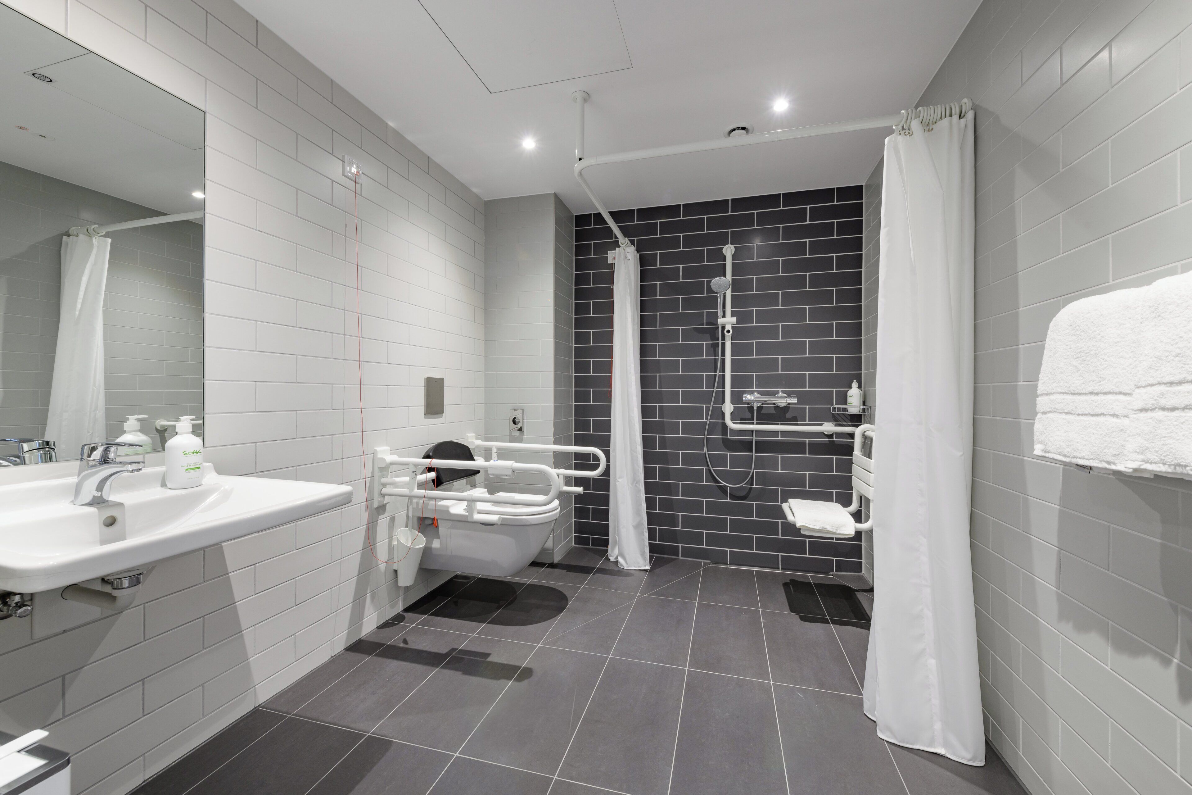 A modern bathroom with a white sink, a toilet with a safety rail, a shower with a black tiled wall, and a white towel on the right.