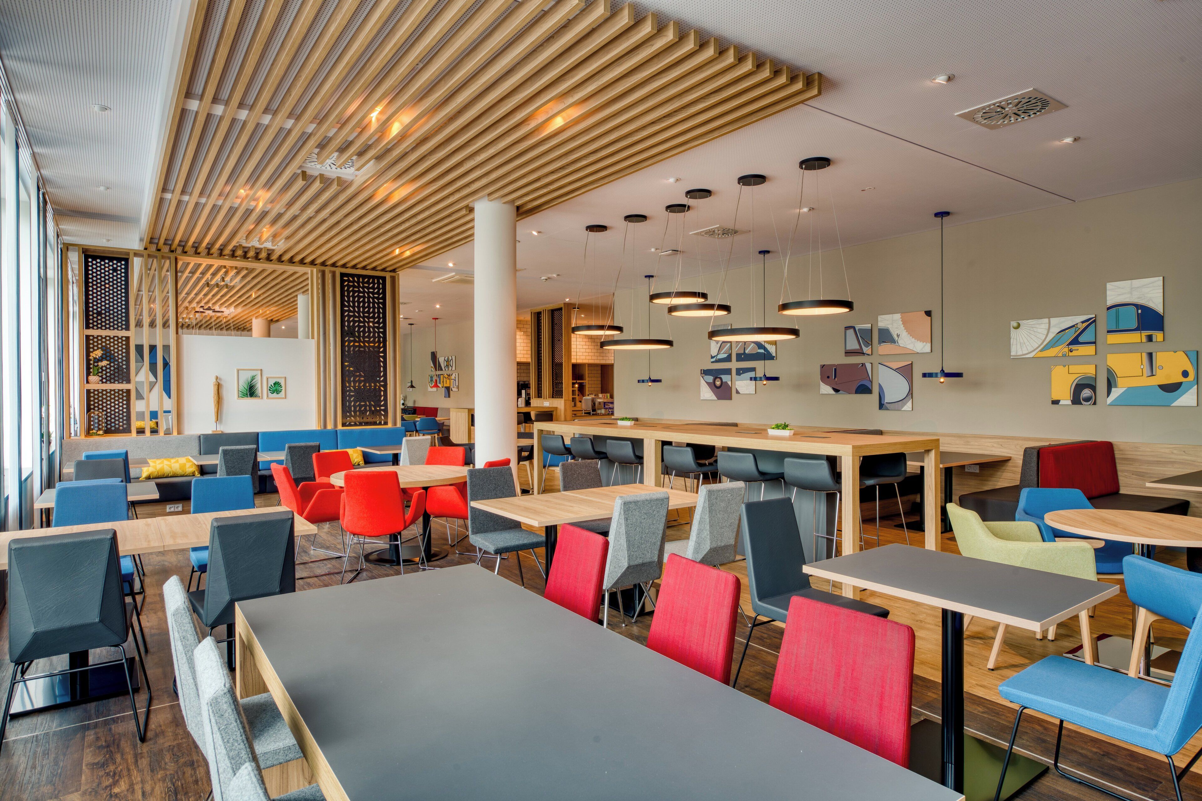 A modern dining area with multiple tables and chairs, wooden ceiling beams, and artwork on the walls.
