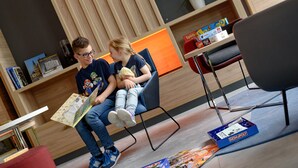 A modern room with a wooden panel wall, a bookshelf with books and a board game, and a red chair. - Holiday Inn Express Fulda by IHG (Fulda)
