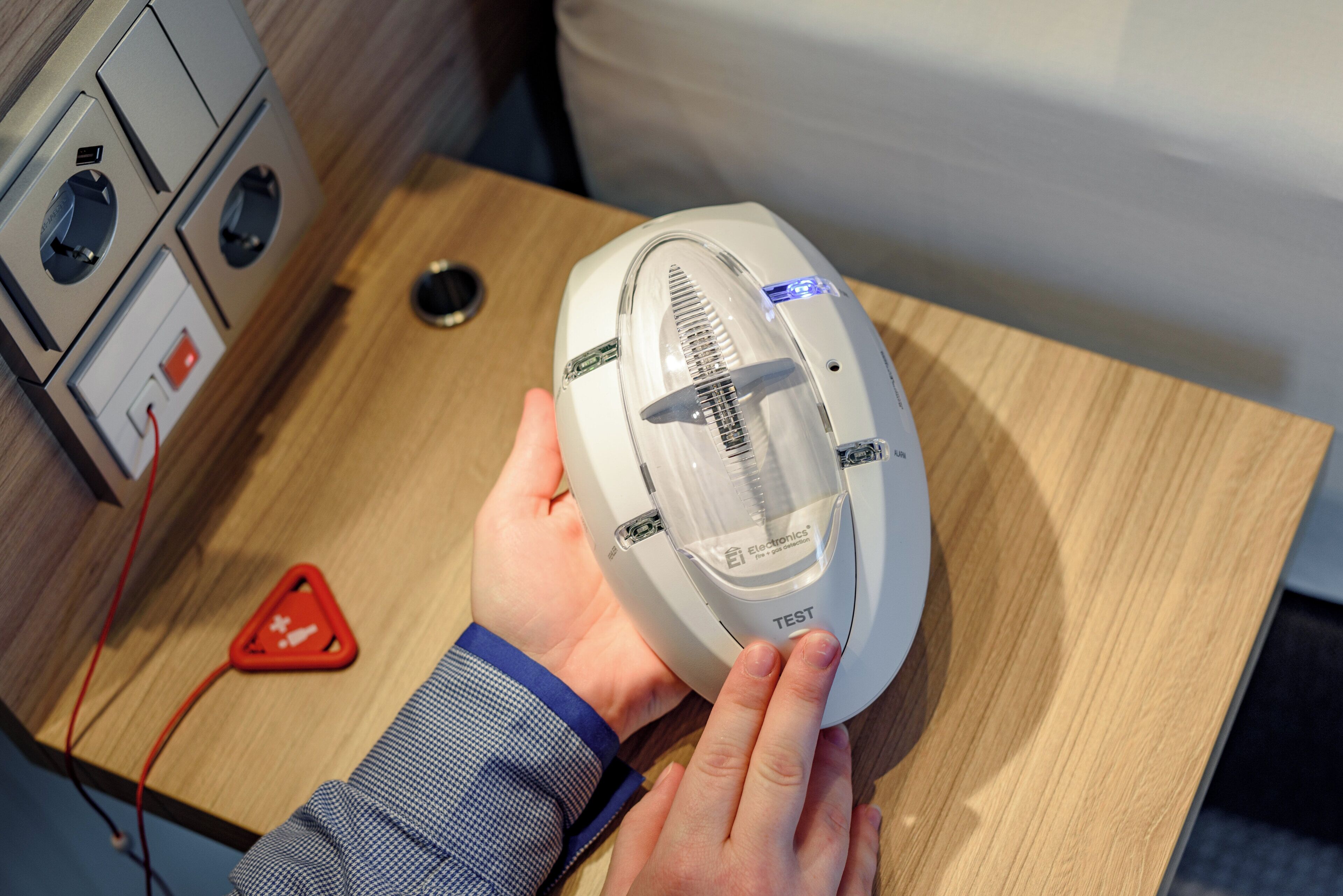 Une personne tient un appareil électronique blanc doté d’un dessus transparent, qui pourrait être un détecteur d’incendie et de gaz, sur une surface en bois.