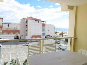A balcony with a table and chairs, overlooking a parking area and buildings with a view of the sea.