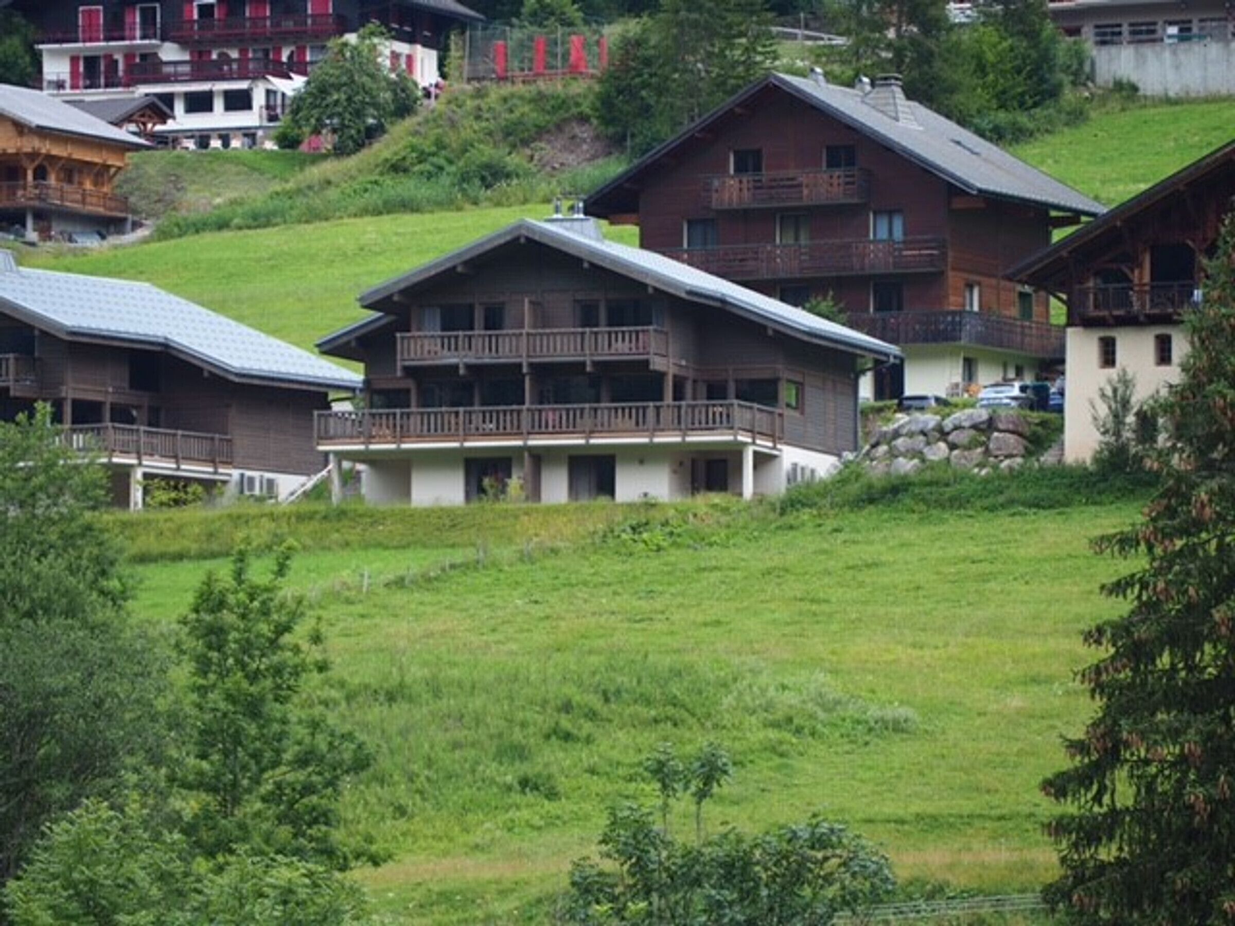 A hillside view of a chalet-style building with a balcony, surrounded by lush greenery and other similar buildings.
