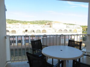 A balcony with a round table and chairs, overlooking a waterfront with buildings and a hill in the background.