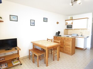 A compact living space with a wooden dining table, chairs, a TV on a stand, a kitchen area with a microwave, and a small cabinet.