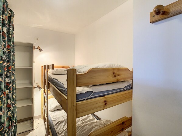 Una cama litera con estructura de madera y ropa de cama blanca, un pequeño estante y una cortina con motivos tropicales.