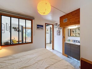 A bedroom with a large bed, a window with blinds, a ceiling light fixture, and a door leading to a bathroom.
