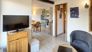 A modern living room with a flat-screen TV, wooden furniture, and a grey sofa. A kitchen is visible in the background with green cabinets and a washing machine. - Modern 2-Bedroom Apartment + Cabin, Close to Combloux Center (Combloux)