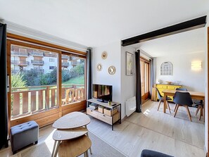 A modern living room with a TV, wooden furniture, a dining area, and a view of a residential building.