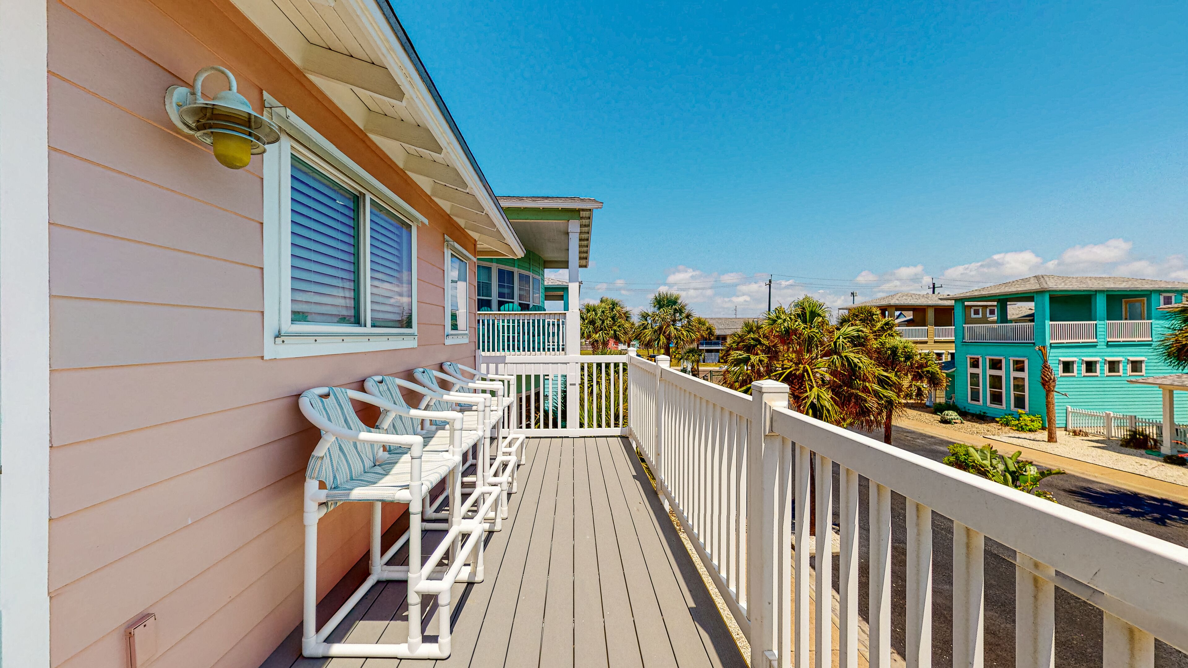 A balcony with white railings and chairs, overlooking a beach with palm trees and turquoise buildings.