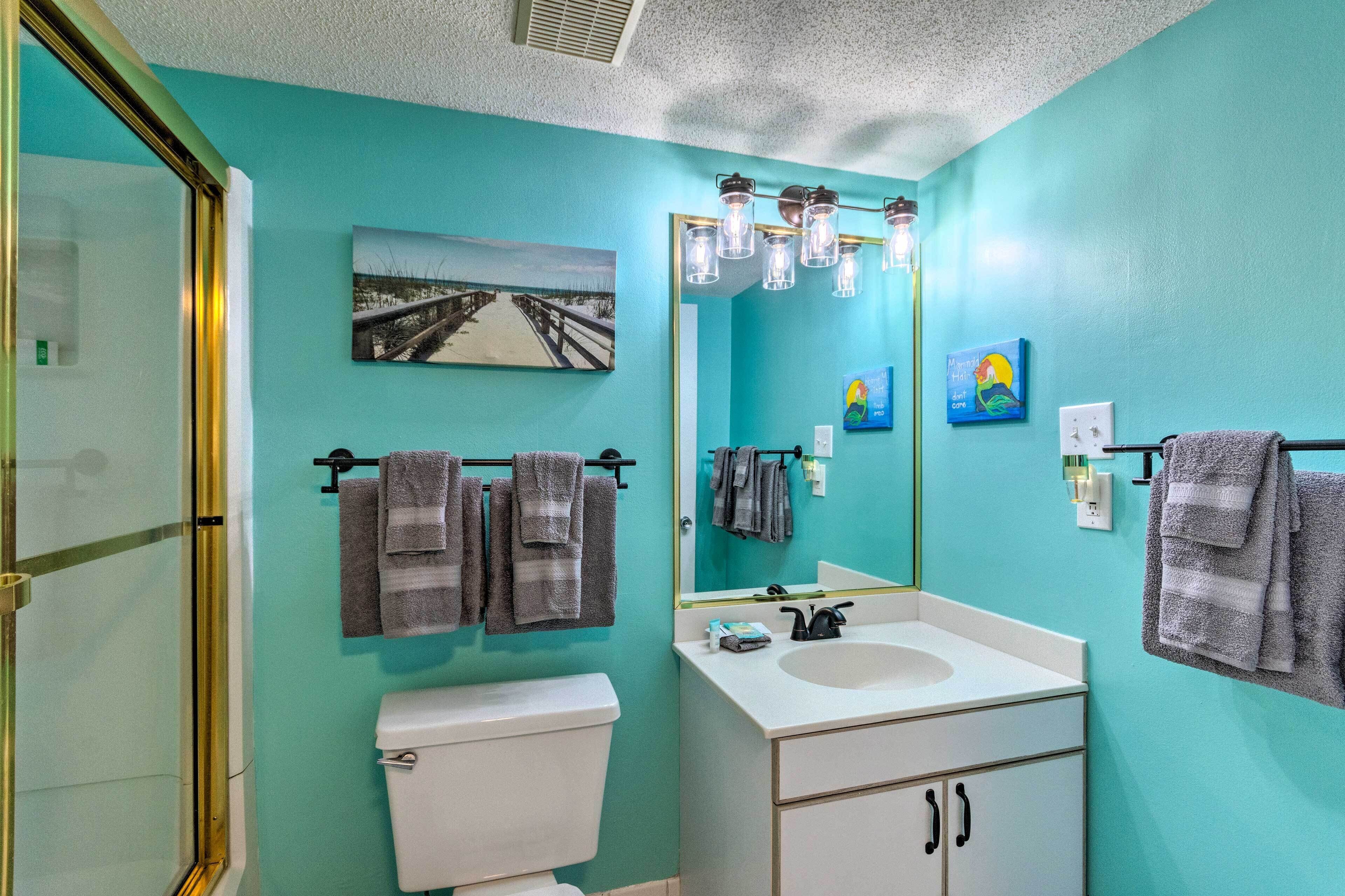 Een badkamer met een wit wastafel, een spiegel en een toilet. Handdoeken hangen aan een rek en er hangt een foto van een strand aan de muur.
