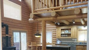 A log cabin interior with a wooden staircase, a fireplace, a dining area, and a kitchen.