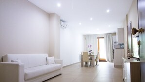 A modern living room with a white sofa, a dining area with a table and chairs, and a kitchenette with a refrigerator and microwave. - Appartamento con Camera da Letto e Balcone (Catania)