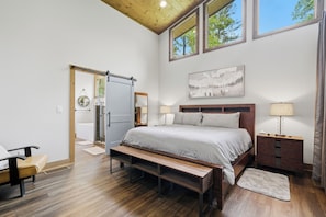 Une chambre à coucher avec un grand lit, des tables de chevet, un banc, un plancher en bois et une vue sur l’extérieur par les fenêtres.