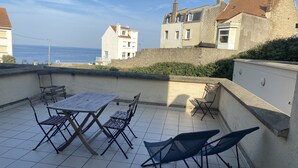 A terrace with a table and chairs, a view of the sea, and buildings in the background.