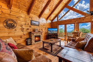 Un interior de cabaña acogedor, con chimenea de piedra, paredes de madera, un televisor y un balcón con vistas.