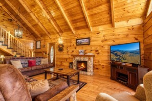 Interior de una cabaña de troncos con chimenea de piedra, televisor, sofá y escalera.