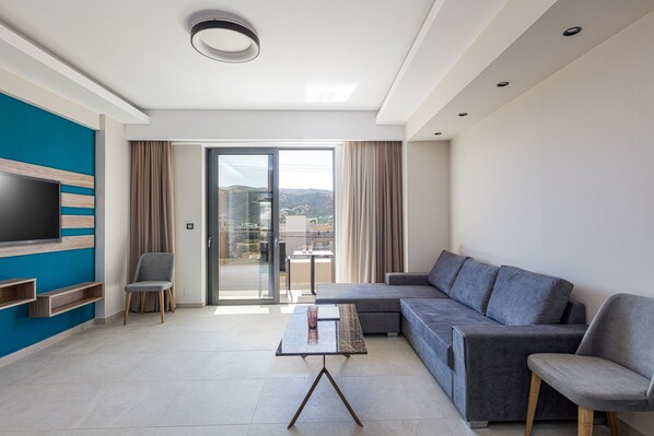 A modern living room with a sectional sofa, a flat-screen TV mounted on a blue wall, a glass door leading to a balcony, and a small table with a vase. - Sapphire Blue Suites (Kissamos)