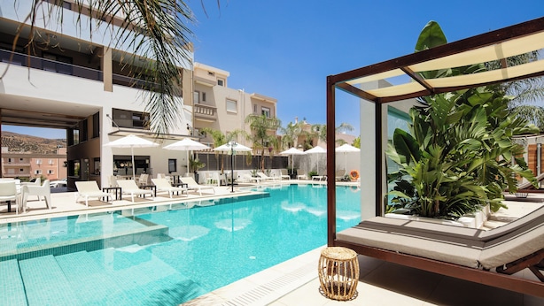 A swimming pool with lounge chairs and umbrellas, surrounded by palm trees and a modern building. - Sapphire Blue Suites (Kissamos)