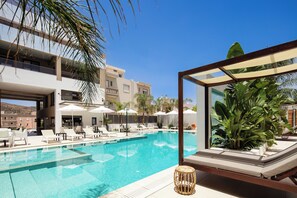 Una piscina con sillas de descanso y sombrillas, rodeada de palmeras y un edificio moderno.