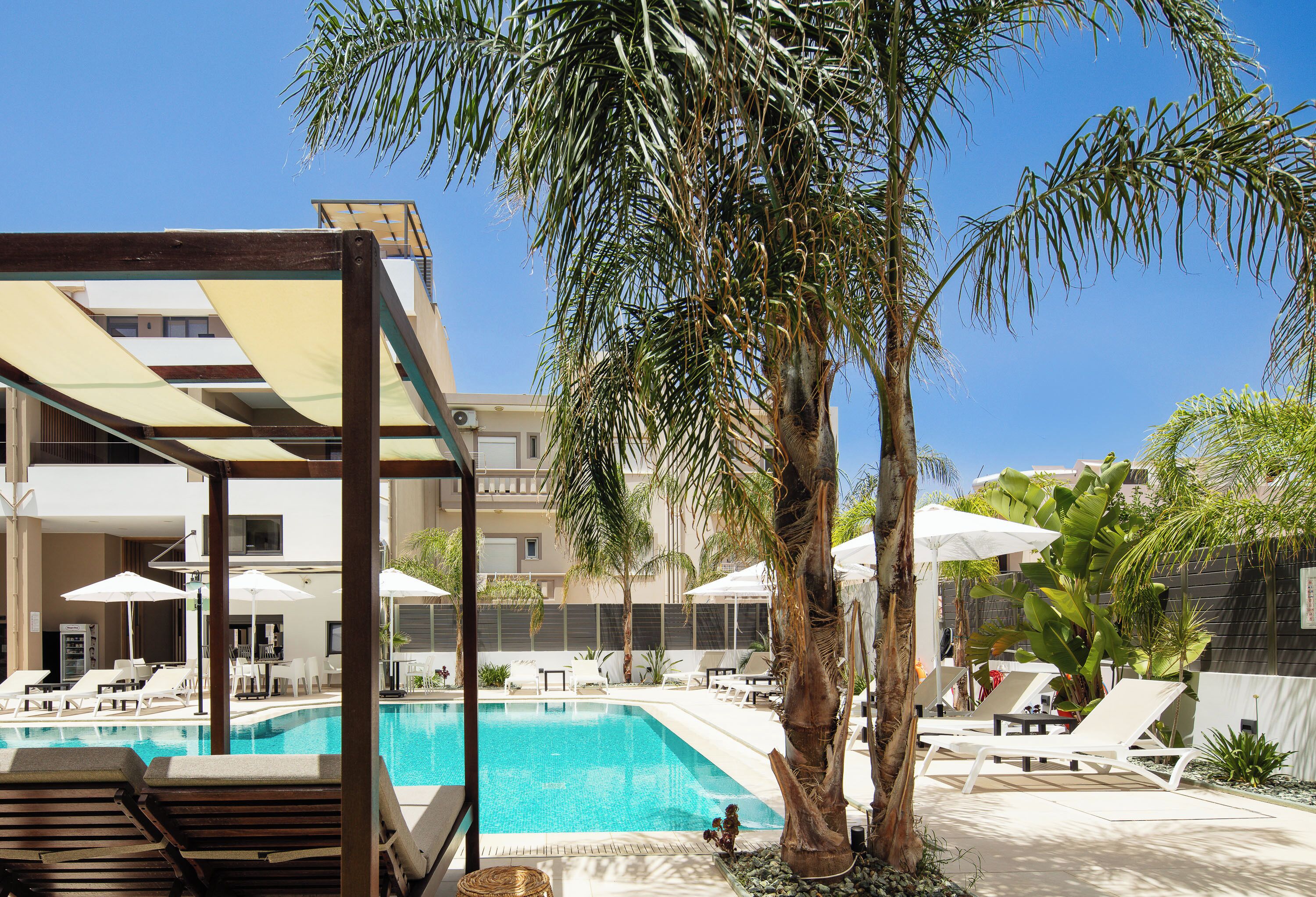 A swimming pool with lounge chairs and umbrellas, surrounded by palm trees and a building in the background.