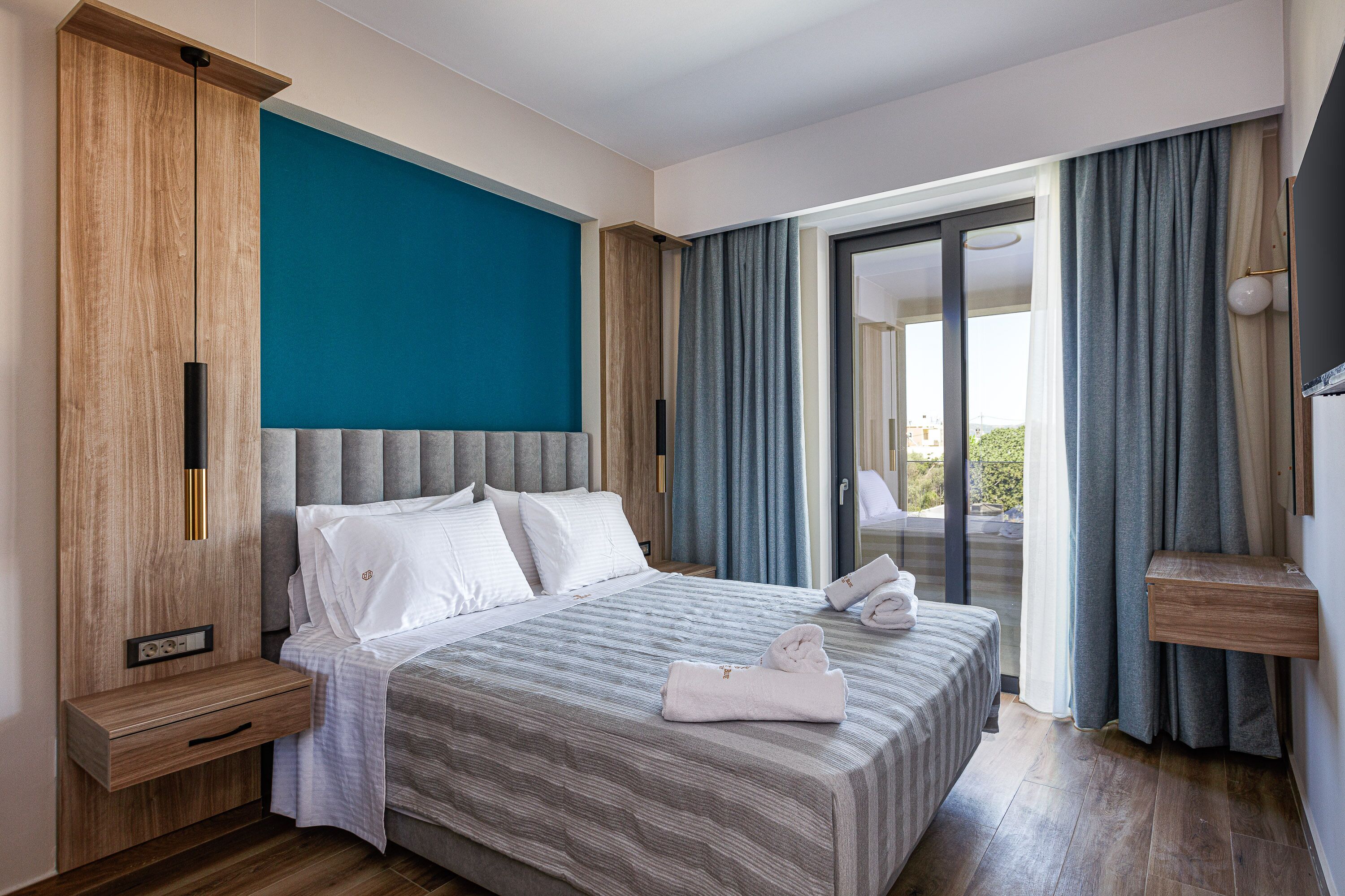 A modern hotel room with a large bed, a wooden nightstand, a built-in shelf, and a view of the outdoors through a sliding glass door.