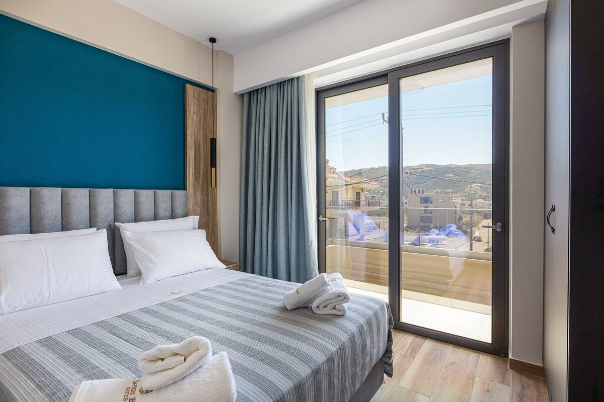 A hotel room with a large bed, a view through a sliding glass door to a balcony, and a blue accent wall.