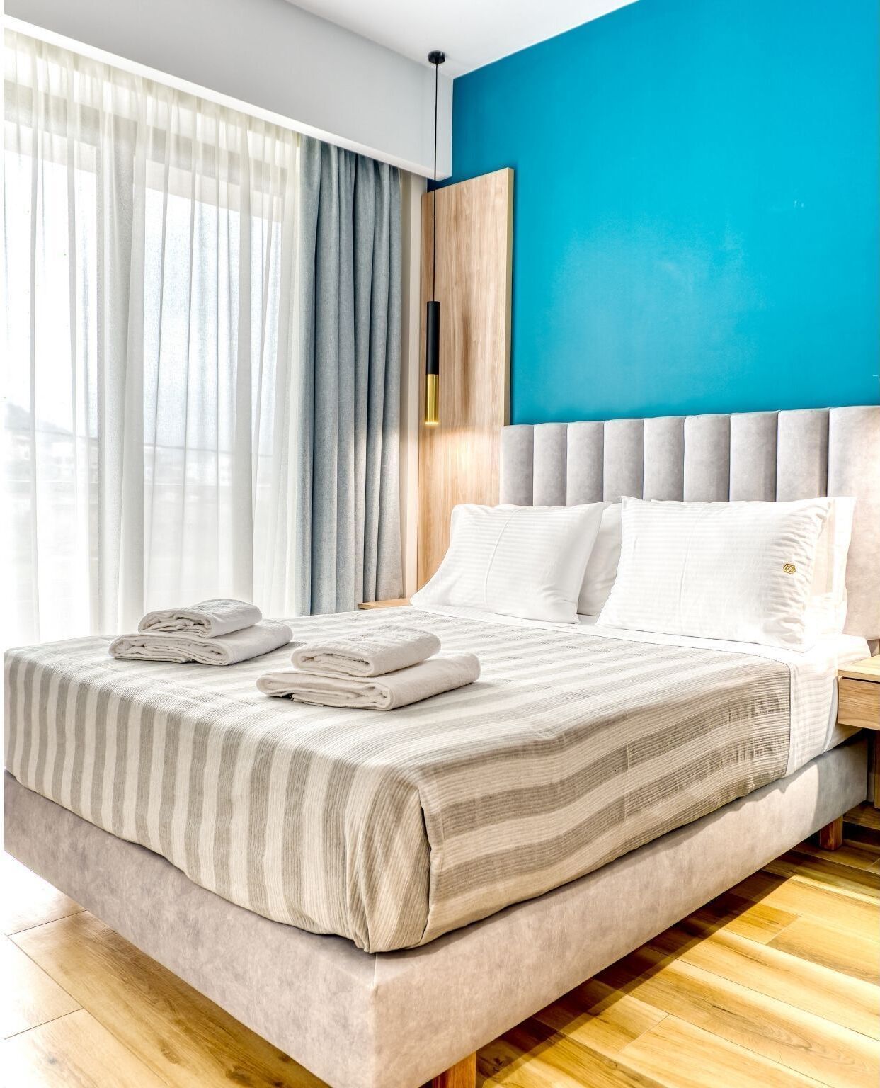 A modern hotel room with a large bed, a wooden headboard, and a view of the city through sheer curtains.