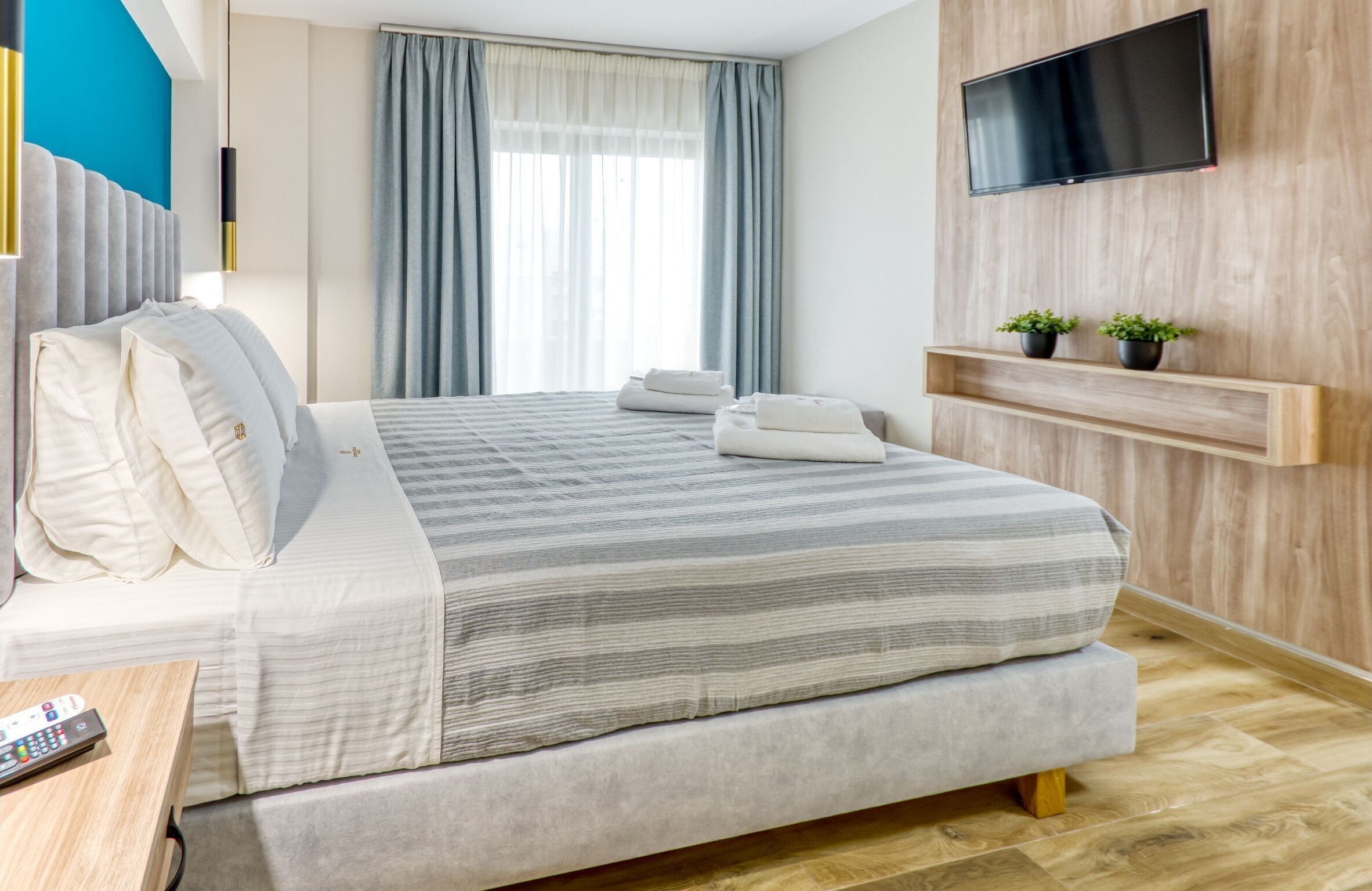 A modern hotel room with a large bed, a TV mounted on the wall, and a wooden shelf with plants.